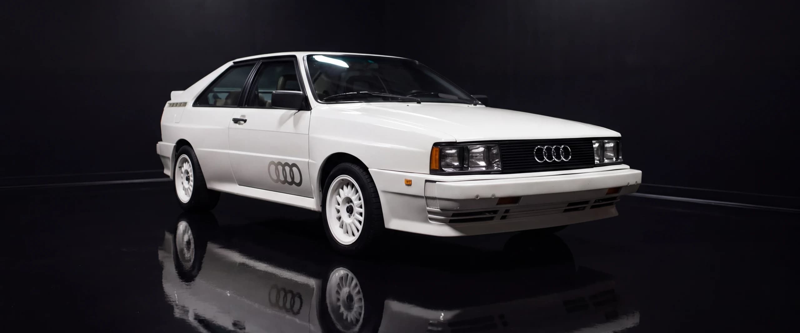 A restored white Audi Quattro lit like a museum exhibit on a mirror-black floor