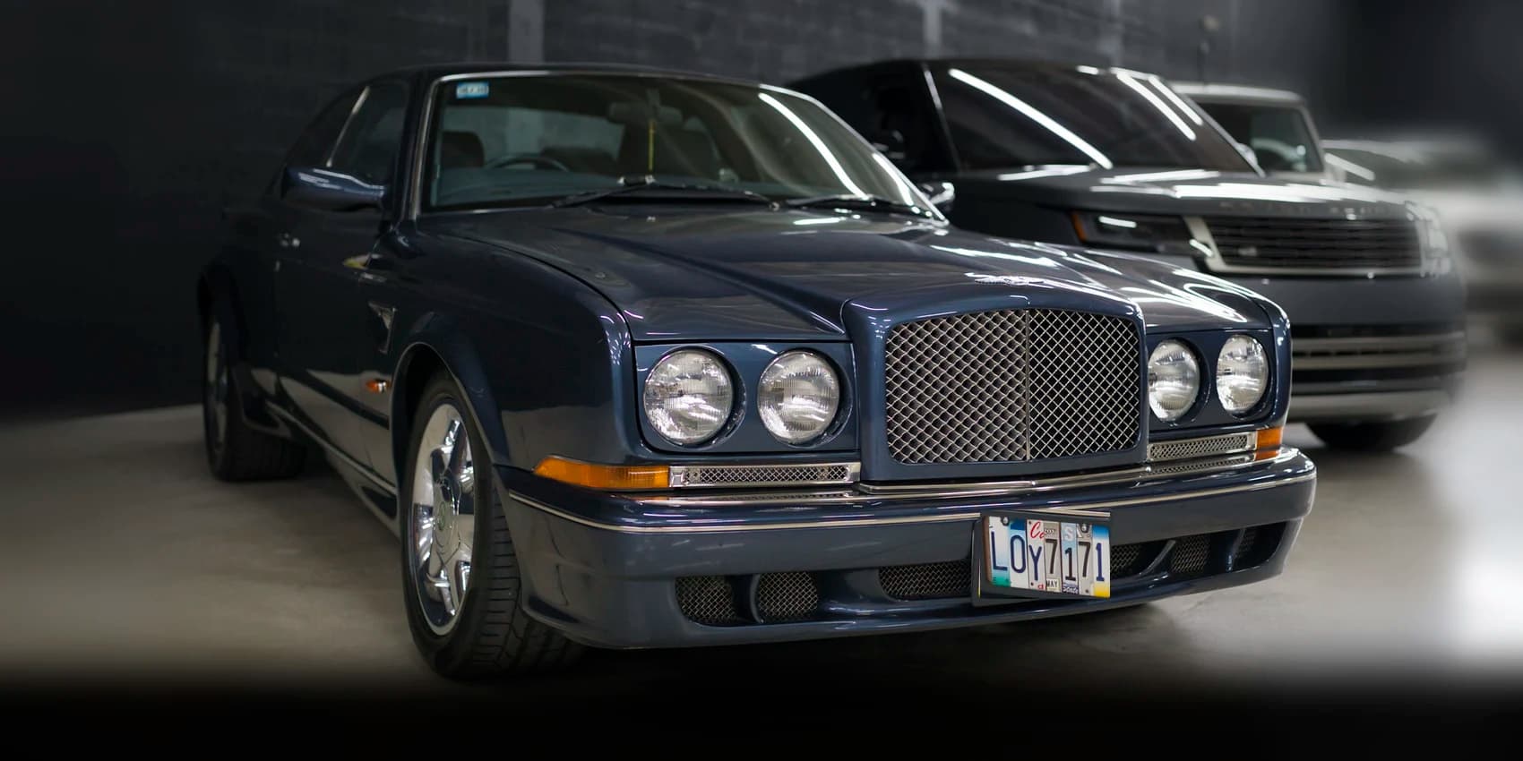 A dark blue Bentley coupe at the head of a row of stored cars, on a dark garage floor