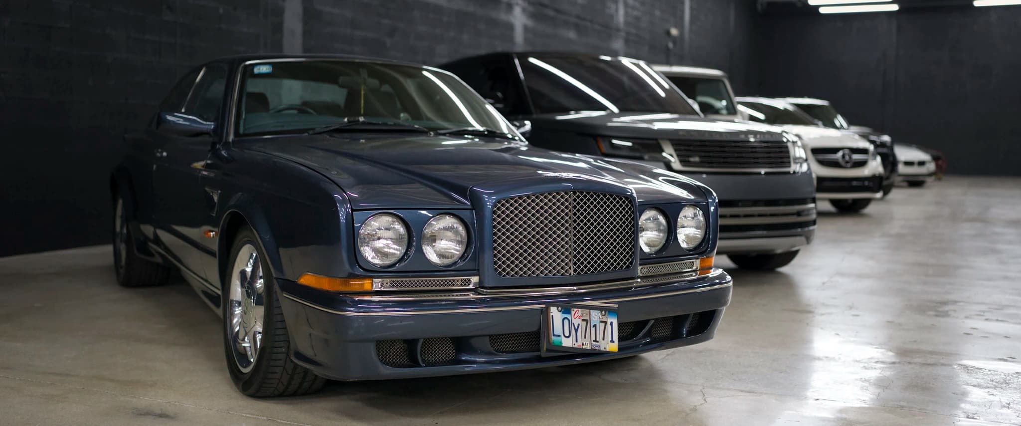 A dark blue Bentley coupe at the head of a row of stored cars on the garage floor