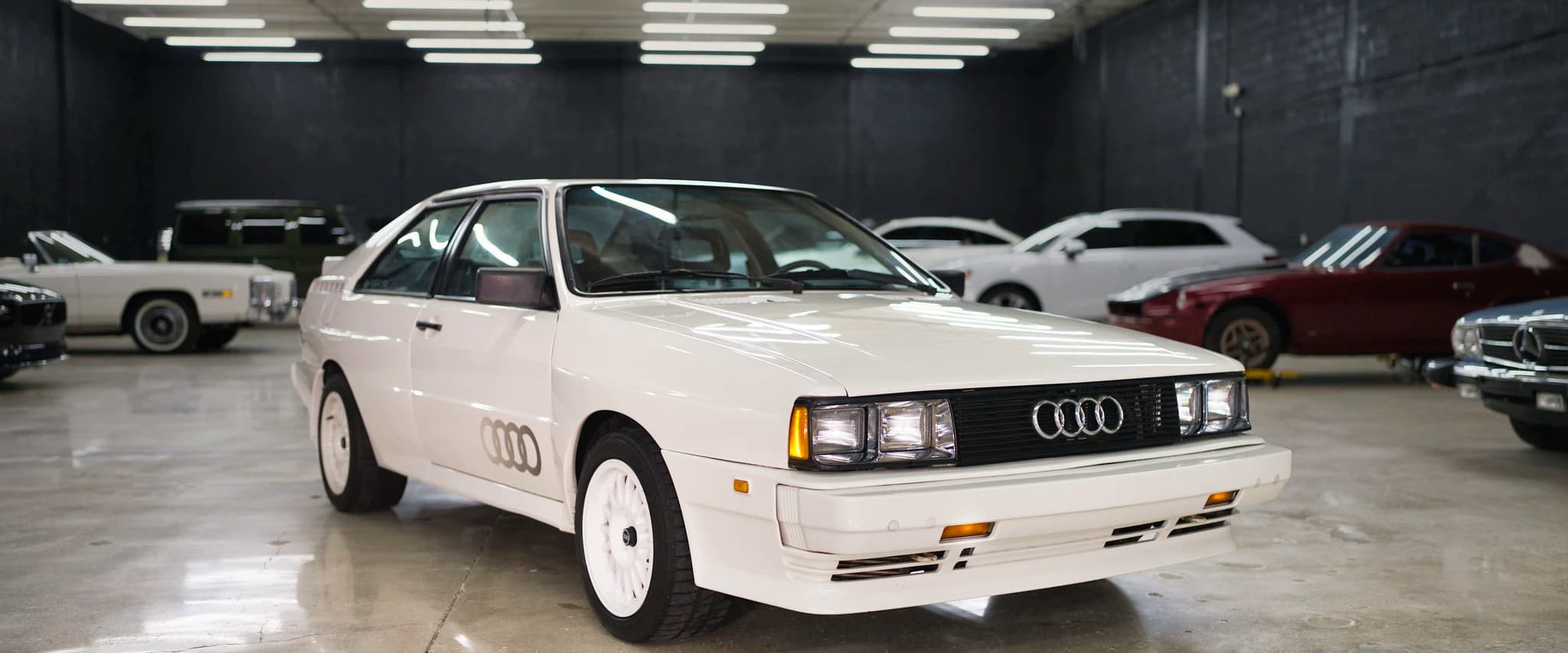 A white Audi Quattro parked on the floor at Full Stop Garage, with stored cars along the wall behind it