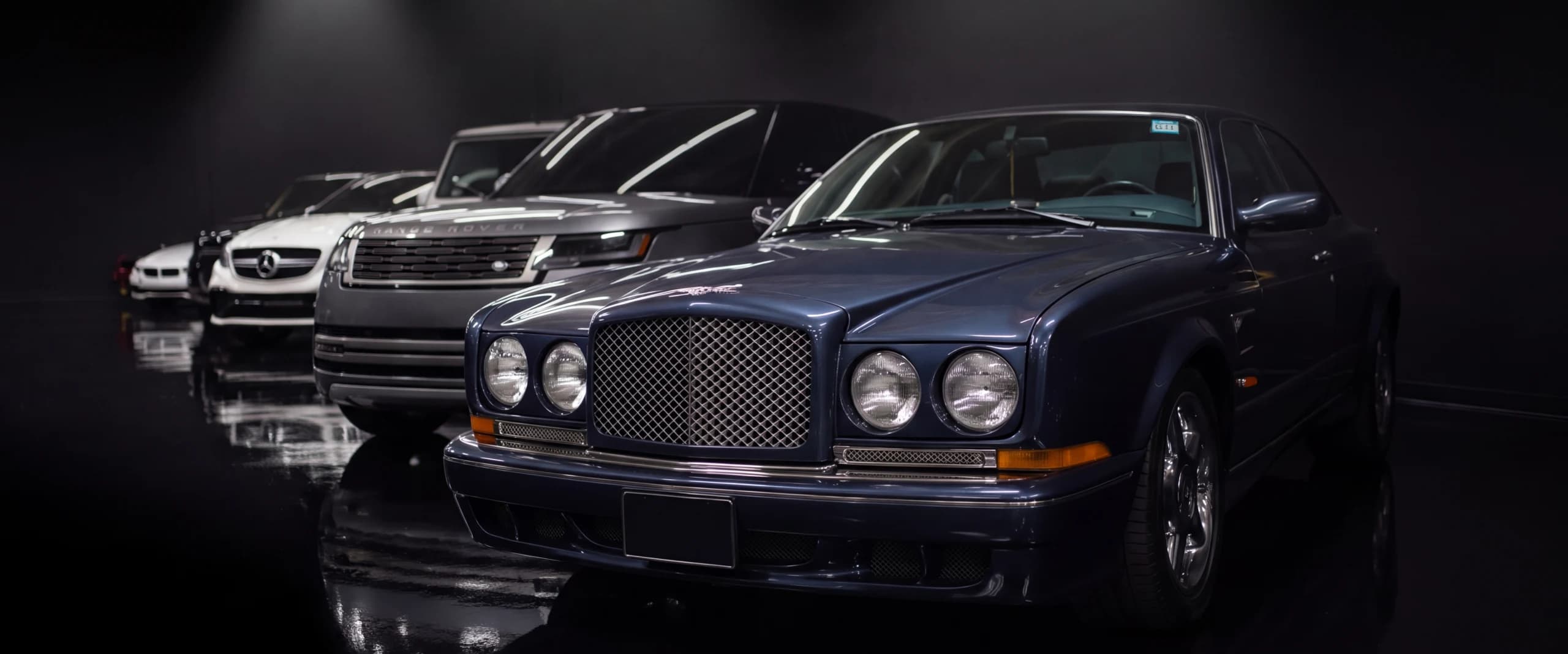 A row of stored cars, a Bentley at the head, each under its own spotlight on a mirror-black floor